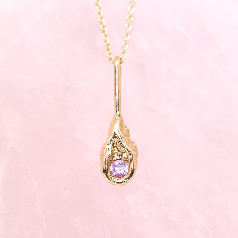 14k yellow gold pendant with a pink sapphire handmade in Toronto. Photographed on a rose quartz background. 