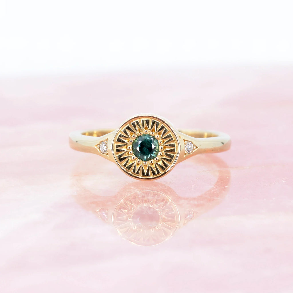 A fancy yellow gold signet ring with hand engraved details and a central teal sapphire is photographed on a rose quartz background.