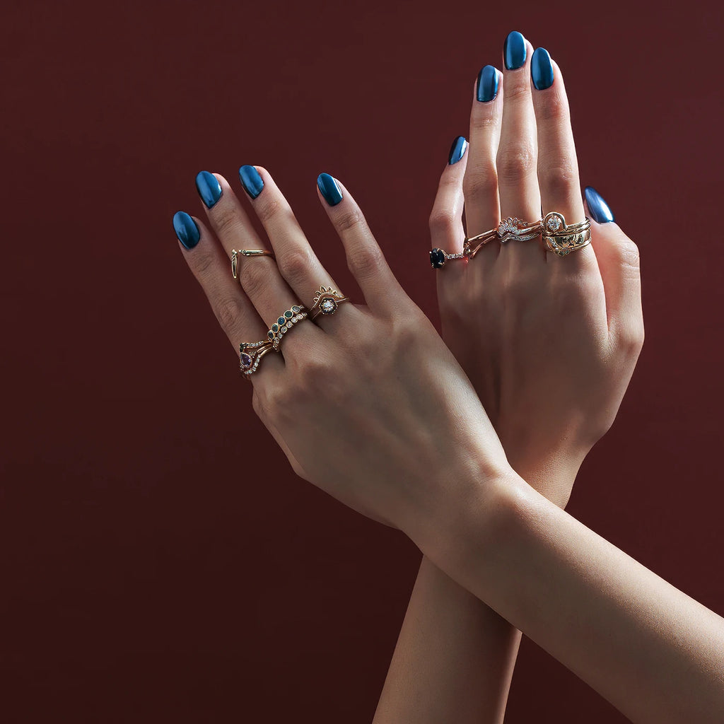 Two hands with metallic blue nails are photographed over a dark red background and wear 17 designer engagement rings and wedding bands featuring diamonds and sapphires. All the jewelry designers of these pieces are Canadian and are represented by independent jewelry store Ruby Mardi.