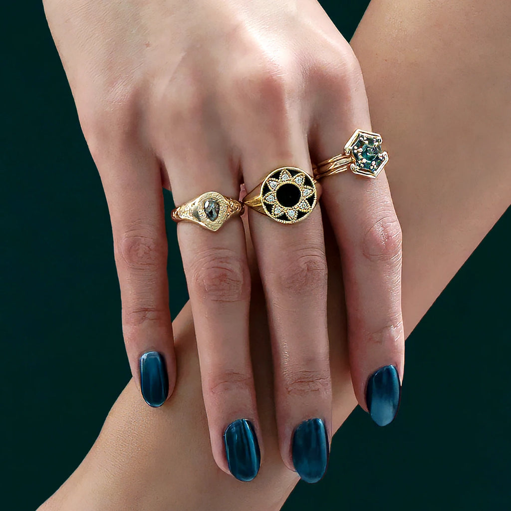 A hand rests on an arm, in a close-up. The hand has metallic blue nails and wears 3 unique and original 18-carat yellow gold rings. One is a signet ring with hand-engraved flowers and a central salt-and-pepper pear-shaped diamond. On the middle finger, a star shape is created with black enamel, natural diamonds and miligrain details. The third ring is a 2-in-1 jewel featuring 13 Australian teal sapphires. The wedding band fits into the engagement ring to create a single ring. Jewelry by Canadian designers. 