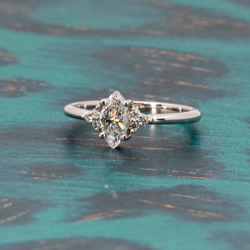 White gold ring with a central lab grown diamond of a marquise shape and 6 small accent diamonds on the sides. The engagement ring is seen on a teal background. 