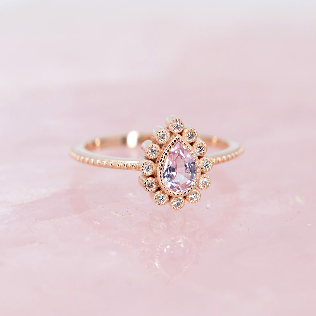 Independent designer ring available immediately at Ruby Mardi jewelry store in Montreal, made in pink gold with natural gems by jewelry designer Emily Gill, this jewel is made with a pear-shaped pink natural sapphire and splendid small round diamonds, seen on a pink background.