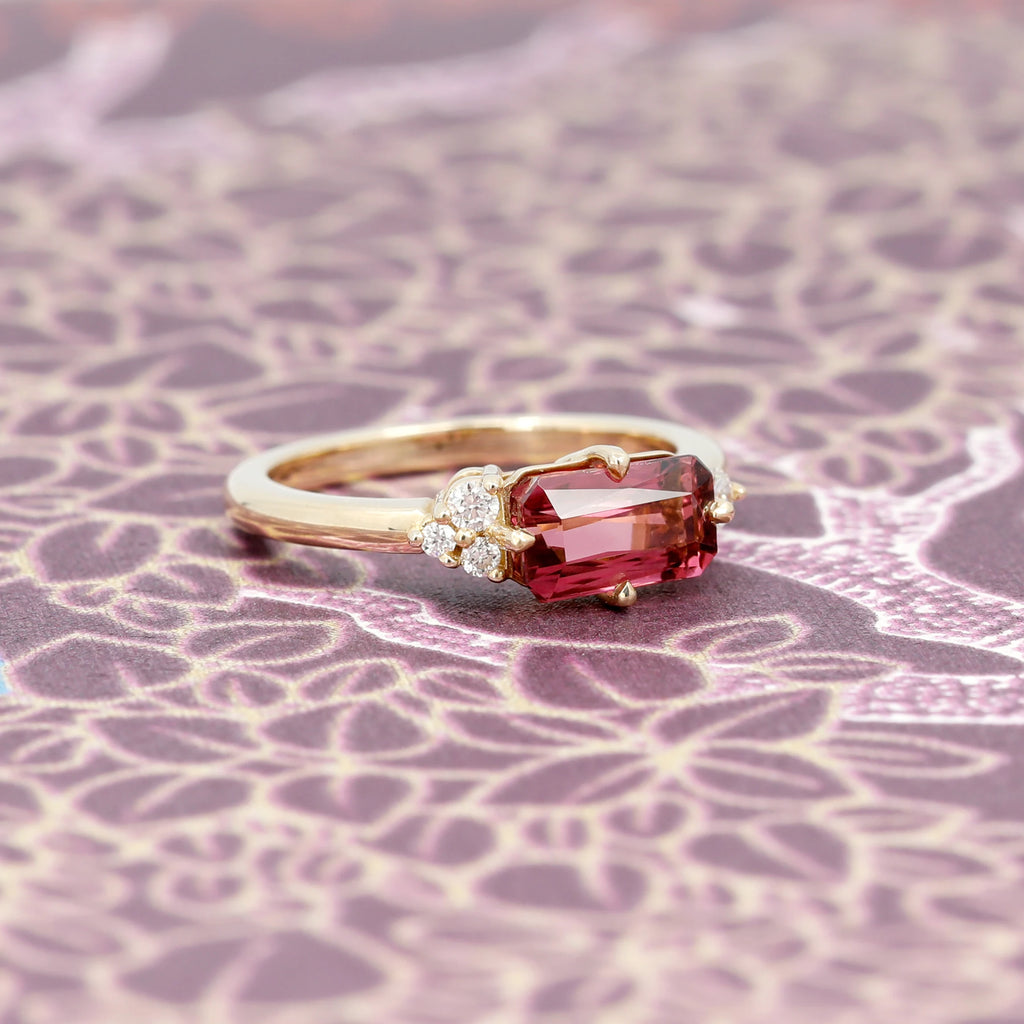 Stunning one-of-a-kind engagement ring featuring a big dark pink tourmaline and asymmetrical diamond accents. This yellow gold bridal ring was designed and handcrafted by indie artist Justine Quintal. It is seen photographed on a flowery background.