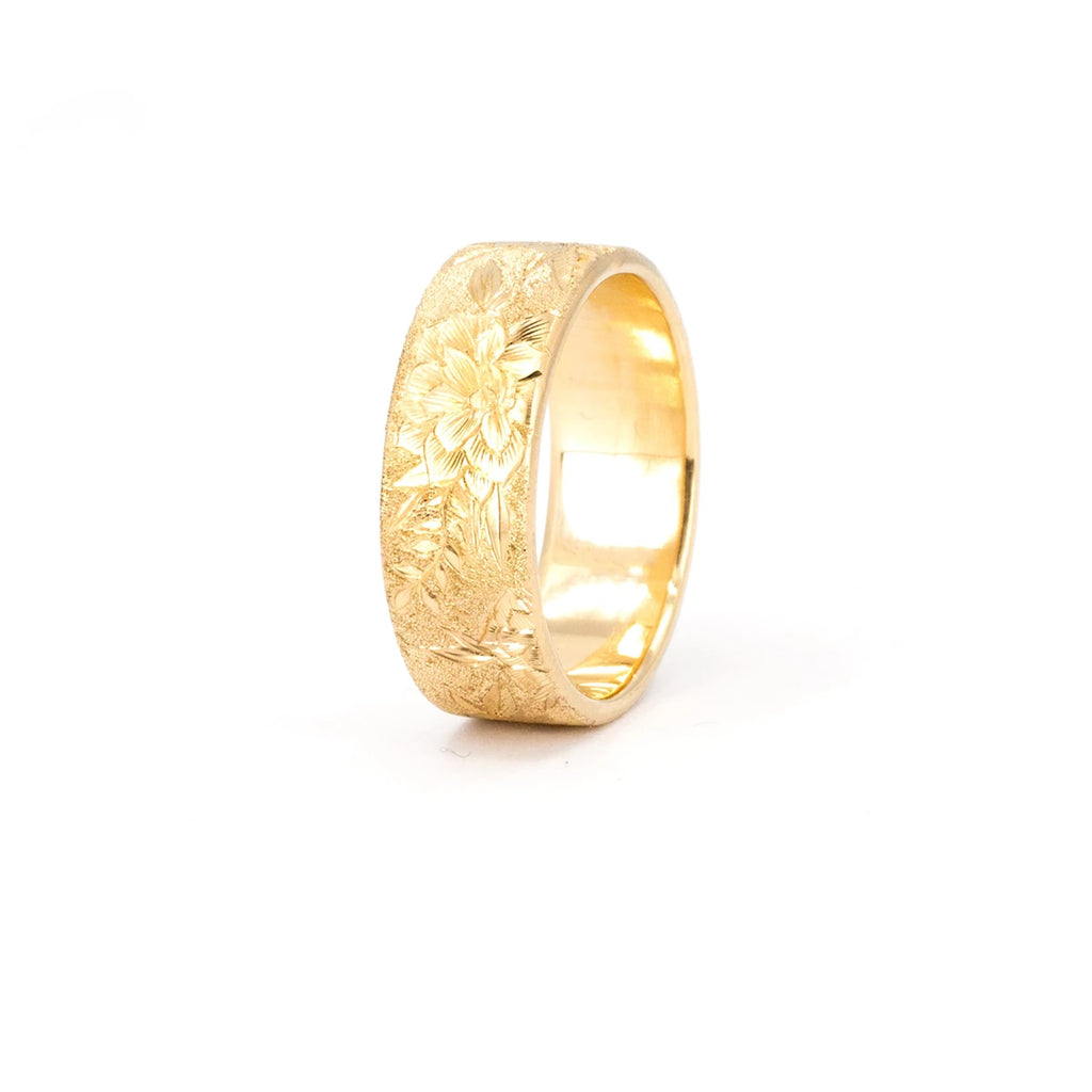 Thick, wide 14-carat yellow gold band featuring finely hand-engraved flowers and a sandblasted texture. The fine jewel is photographed against a white background and is slightly overexposed. The wedding ring was created by independent brand Rebel & Rogue. This is a unique piece of bridal jewelry only available at Ruby Mardi.