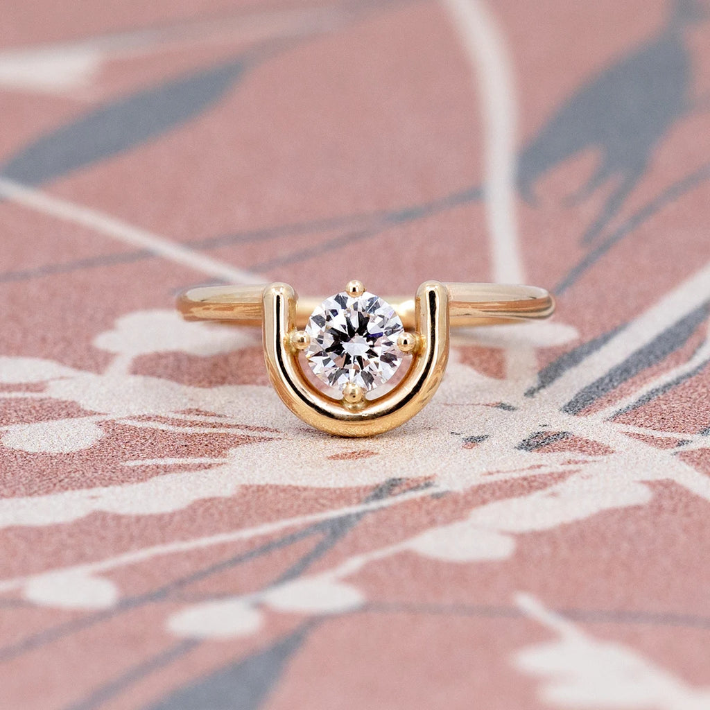 A prong-set, brilliant cut diamond is partially nestled within an arching 14K yellow gold band, one side wrapped in gold while the other side floats free. The ring is seen photographed on a funky background. This is the work from indie Canadian designer Erica Leal.