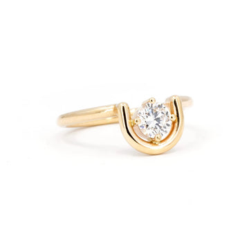 A prong-set, brilliant cut diamond is partially nestled within an arching 14K yellow gold band, one side wrapped in gold while the other side floats free. The ring is seen photographed on a white background. This is the work from indie Canadian designer Erica Leal.