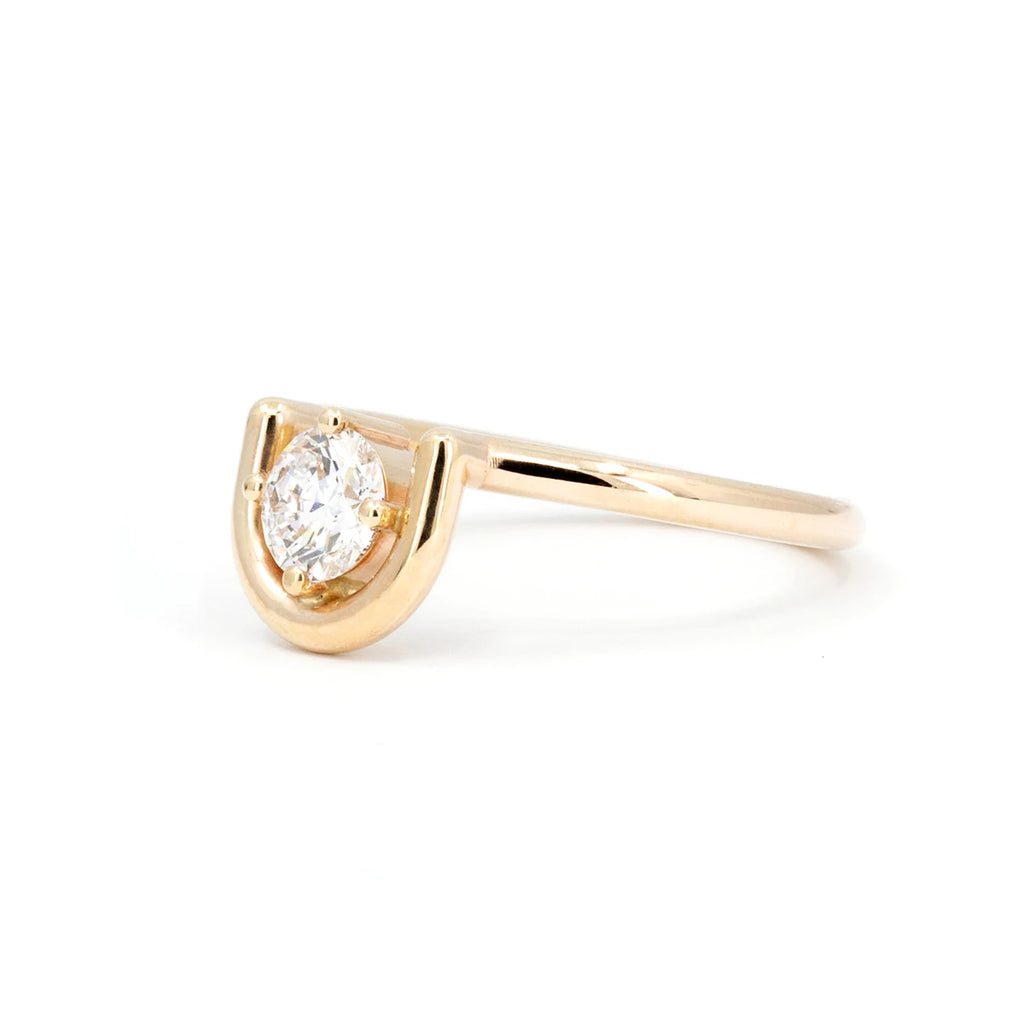 A prong-set, brilliant cut diamond is partially nestled within an arching 14K yellow gold band, one side wrapped in gold while the other side floats free. The ring is seen photographed on a white background and seen from its side. This bridal piece is available at fine jewellery store Ruby Mardi.