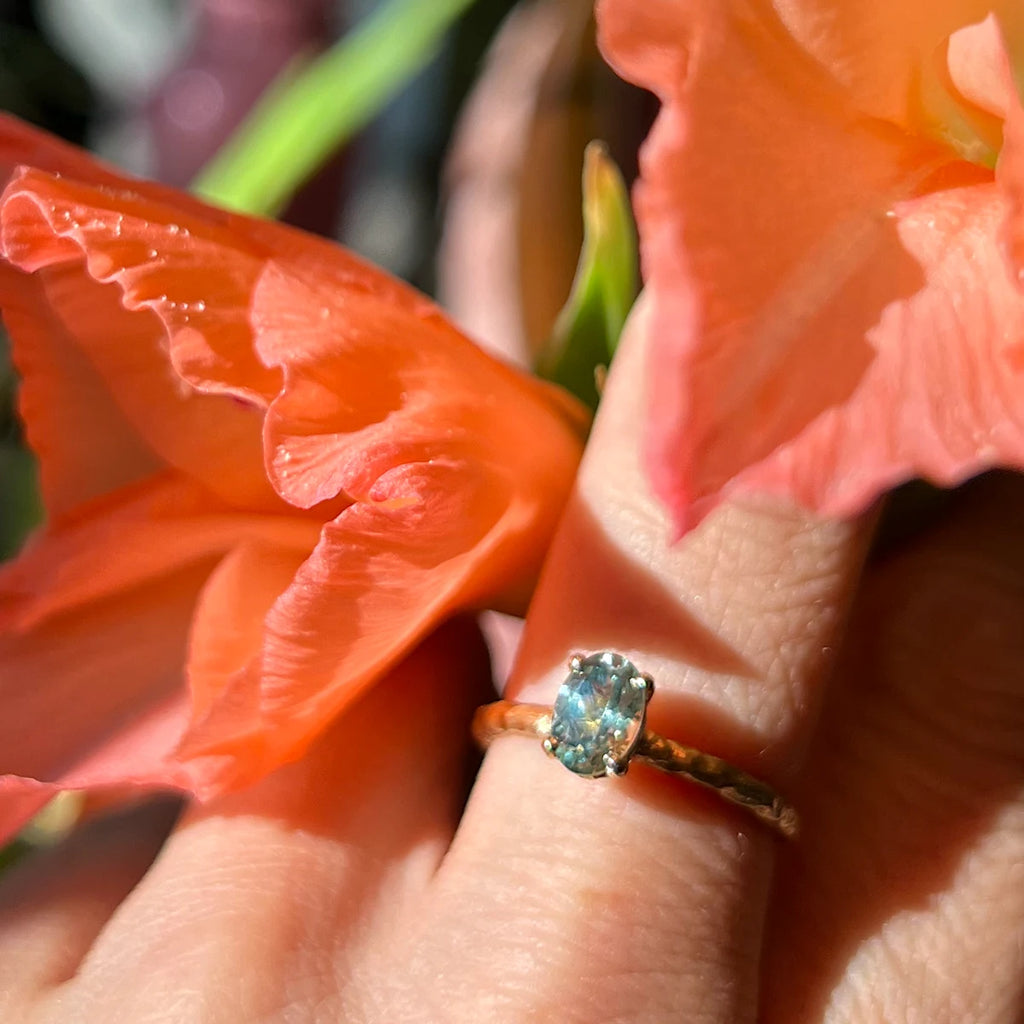 One-of-a-kind ring handmade by Anouk Jewelry seen worn in close up on a hand holding orange flowers. It is photographed in plain sun and the green gemstone shines. 