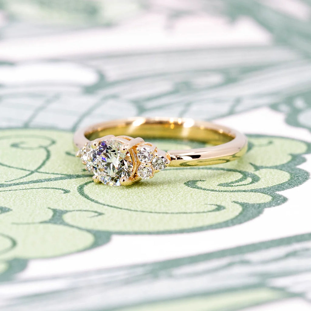 A classic engagement ring in yellow gold with round brilliant diamonds is seen photographed on an abstract green background. This bridal canadian jewelry piece was designed and handcrafted in Montreal by the best jewelry store in Canada, Ruby Mardi.