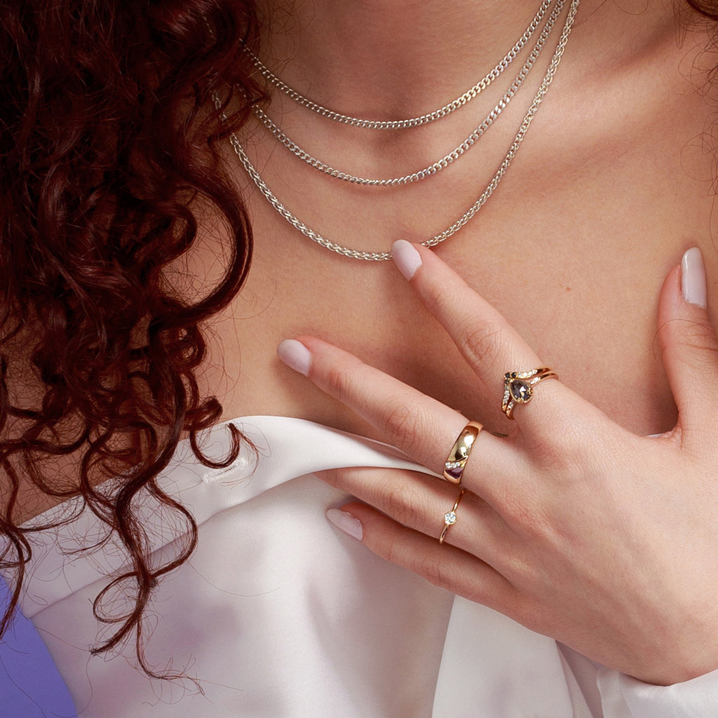  A lady in close up wearing yellow gold rings with Canadian diamonds : solitaire engagement ring, wide wedding band, and a bridal set. She’s also wearing three silver chains. 