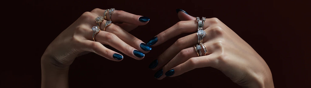 Two hands in close up are filled with designer diamond rings. The background is dark, almost black, and her nails are metallic blue.