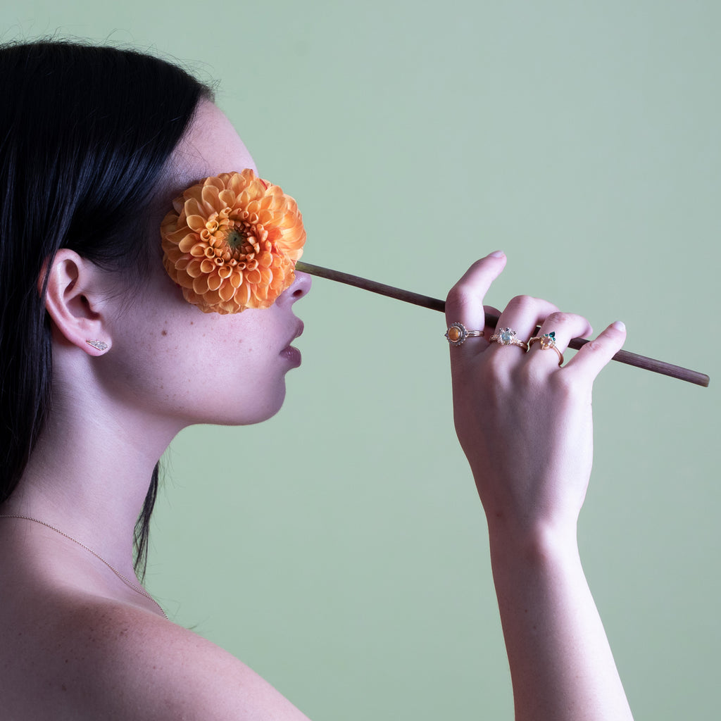 An Asian woman in profile hides her eye with an orange dahlia. On her hand are 4 rings by independent designers, all made in Canada.