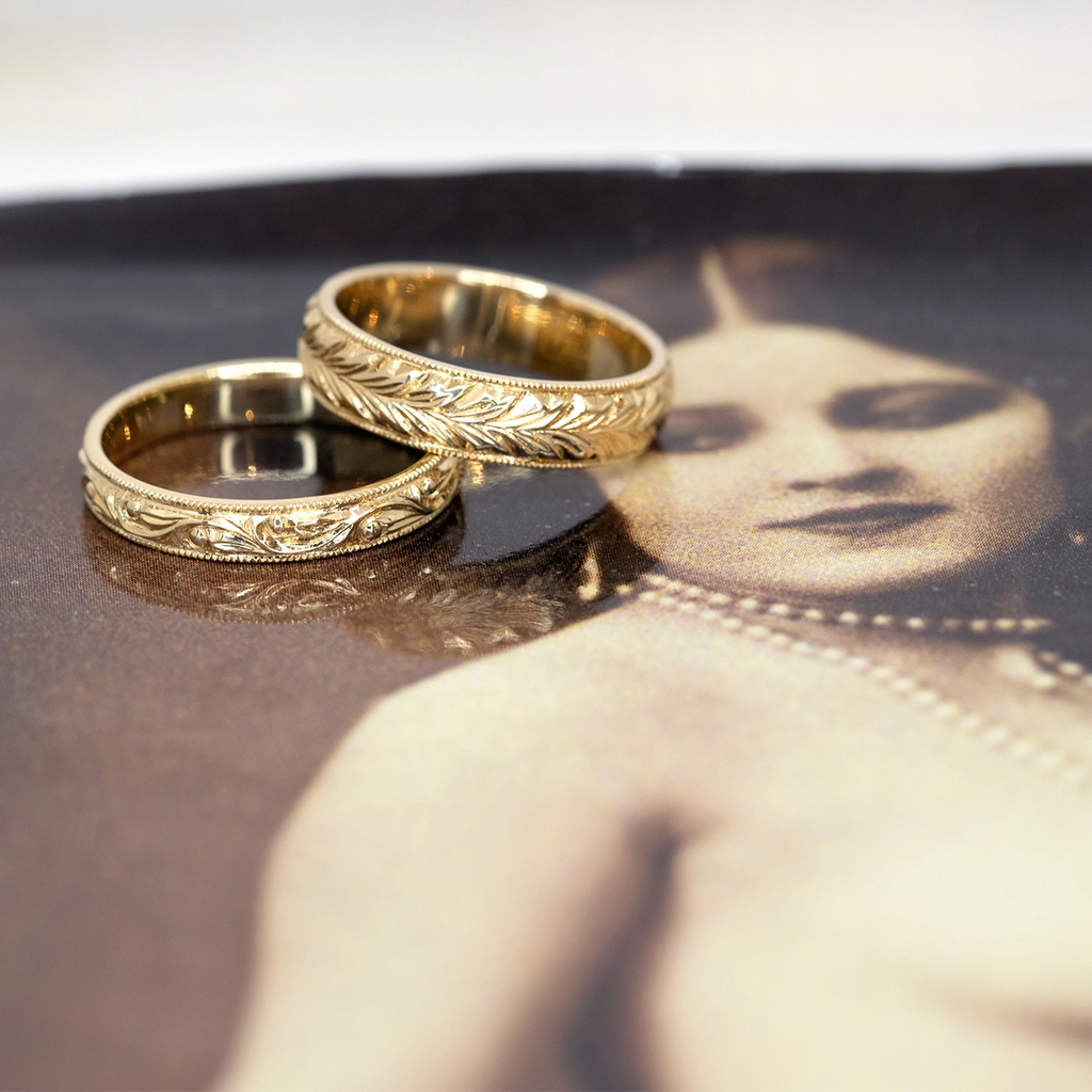 Two hand-engraved yellow gold wedding bands are photographed on an old sepia picture of a lady with a pearl necklace.