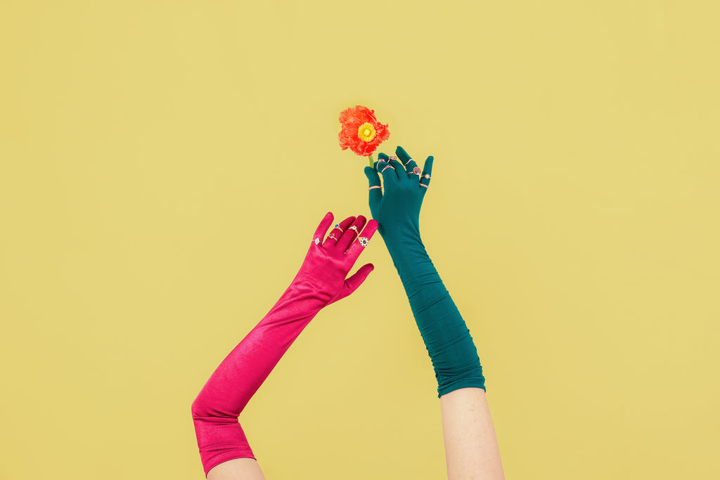 Two hands wearing long, colorful gloves are photographed against a yellow background with a strong contrast. Both hands are wearing several Canadian designer rings.