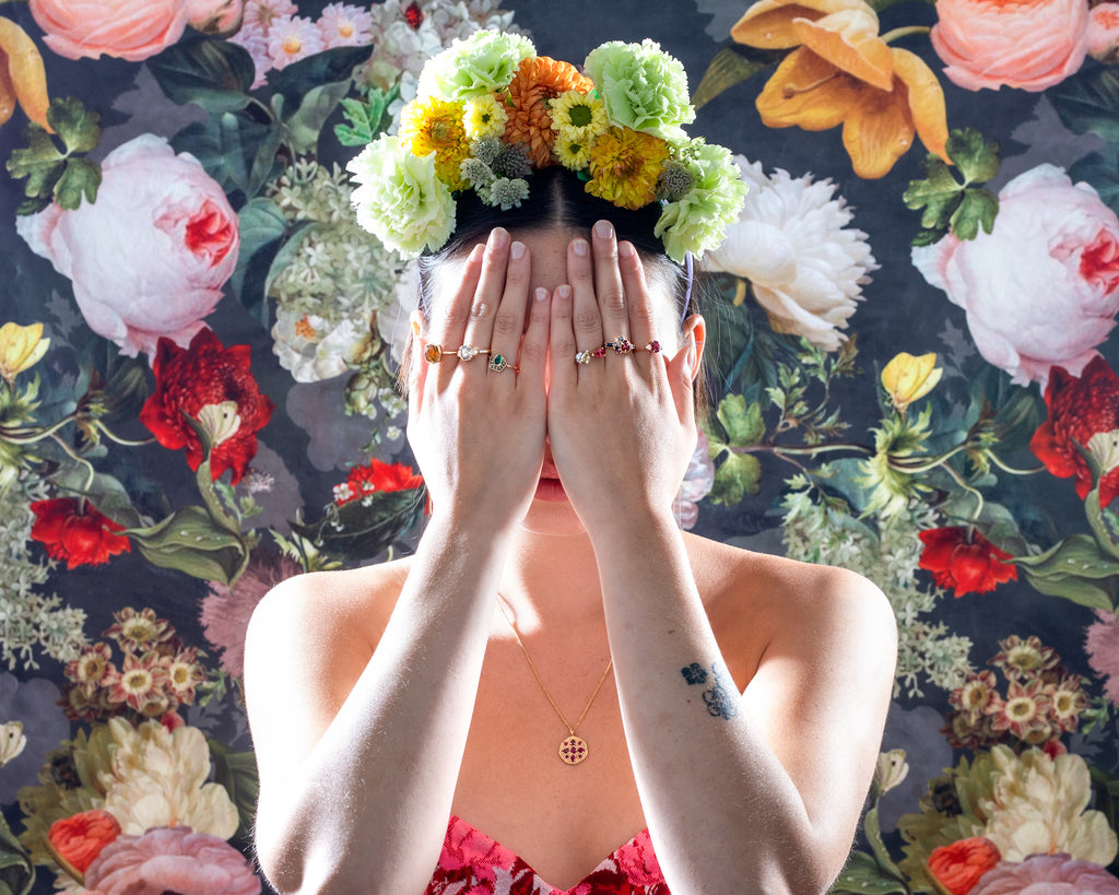 A creative jewelry photoshoot inspired by Frida Kahlo. A young woman wearing a floral bustier and a crown of flowers on her head hides her face with both hands. She's wearing 6 unique, original designer rings.