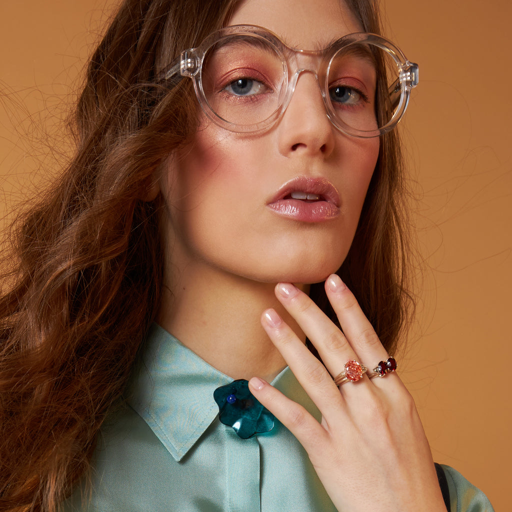 Fashion photo shoot. Young woman photographed close-up wearing large plastic glasses, holding a hand on her chin with two large gemstone rings.