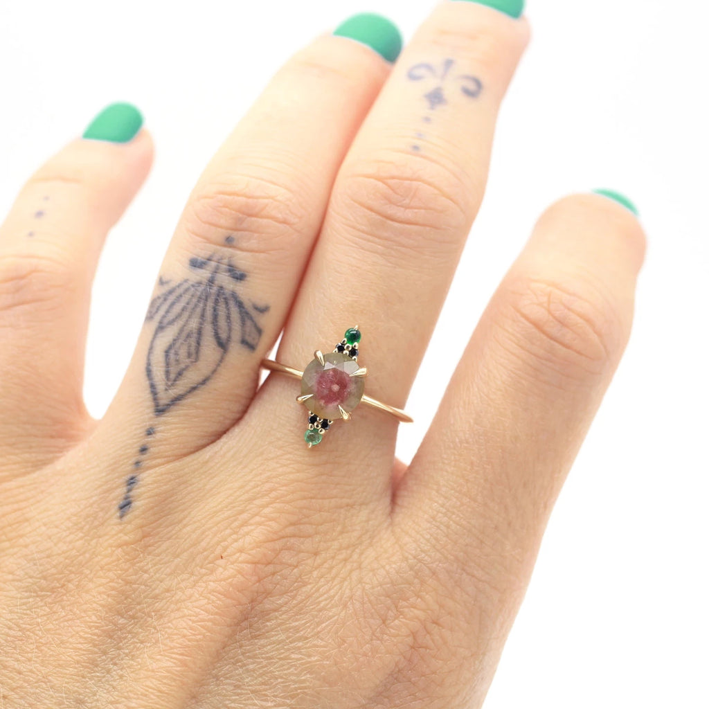A hand wearing an alternative engagement ring is photographed in close up. The hand has a tattoo and turquoise nails, and the ring shows a beautiful watermelon tourmaline, black diamonds and genuine emeralds.