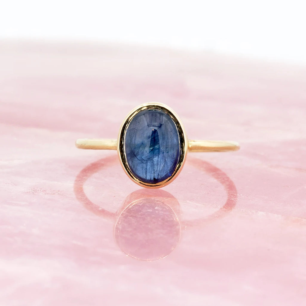 A yellow gold ring featuring a bezel set blue sapphire cabochon is photographed on a quartz rose background. A designer jewelry piece available at Ruby Mardi.