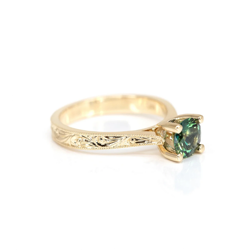 Hand-engraved yellow gold solitaire ring featuring a teal sapphire centerpiece. Intricate floral details on the band, handcrafted by an independent Canadian jeweler. Available at Ruby Mardi.