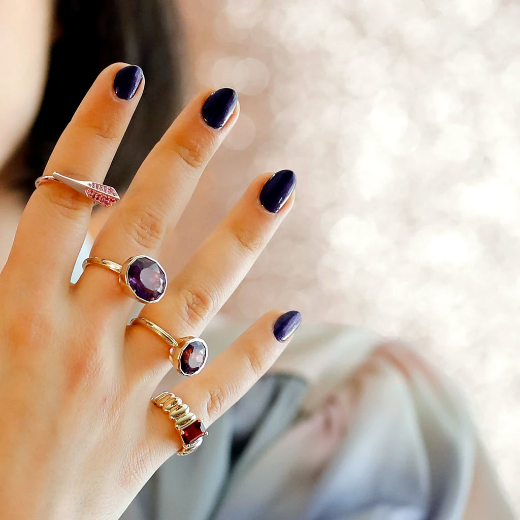 We see half the face of a young woman with red lips and black bobbed hair, out of focus, photographed in front of a pink glittery background. Her hand is in the foreground, in focus, and she's wearing 4 highly original rings by independent designers. Three of the rings are very large and feature colored stones: a large natural ruby cabochon, a large rose quartz and a large lemon quartz. The fourth ring is a slender arrow-shaped ring in yellow gold, set with a diamond pavé.