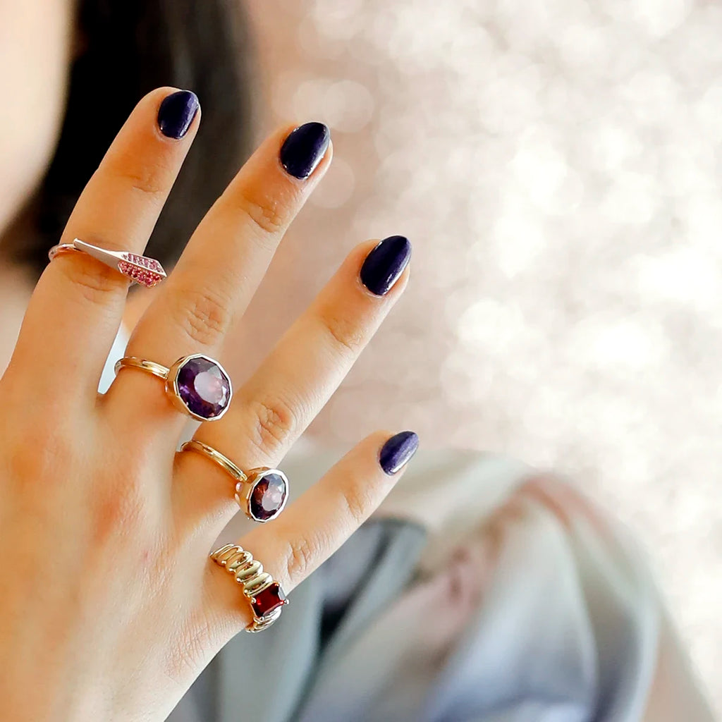 A hand rises to the foreground. Behind it we see a shoulder in a pastel blouse and black bobbed hair. The hand features 4 original and unique designer rings: two very large bezel-set rings with natural amethysts, a large gold ring with a red garnet and a tapering pink gold ring with a pink sapphire pavé.