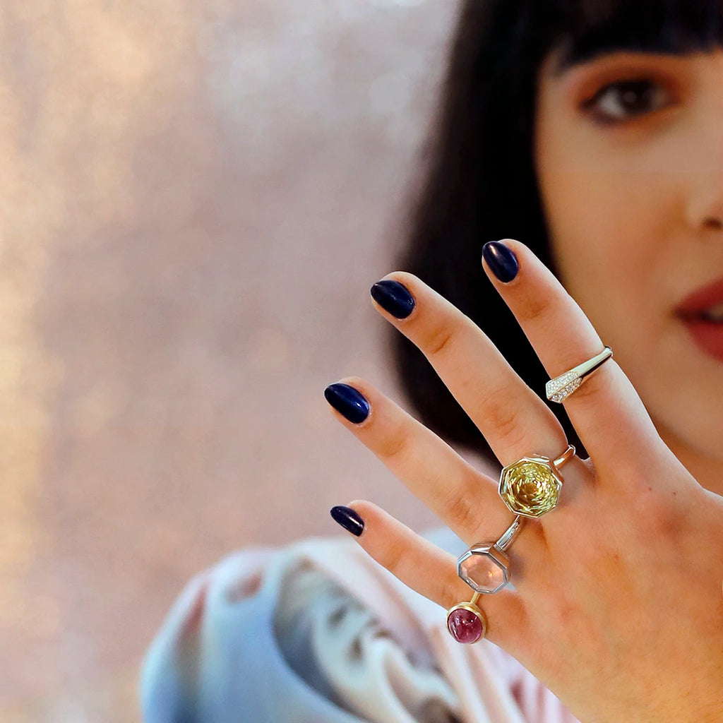 We see half the face of a young woman with red lips and black bobbed hair, out of focus, photographed in front of a pink glittery background. Her hand is in the foreground, in focus, and she's wearing 4 highly original rings by independent designers. Three of the rings are very large and feature colored stones: a large natural ruby cabochon, a large rose quartz and a large lemon quartz. The fourth ring is a slender arrow-shaped ring in yellow gold, set with a diamond pavé. 