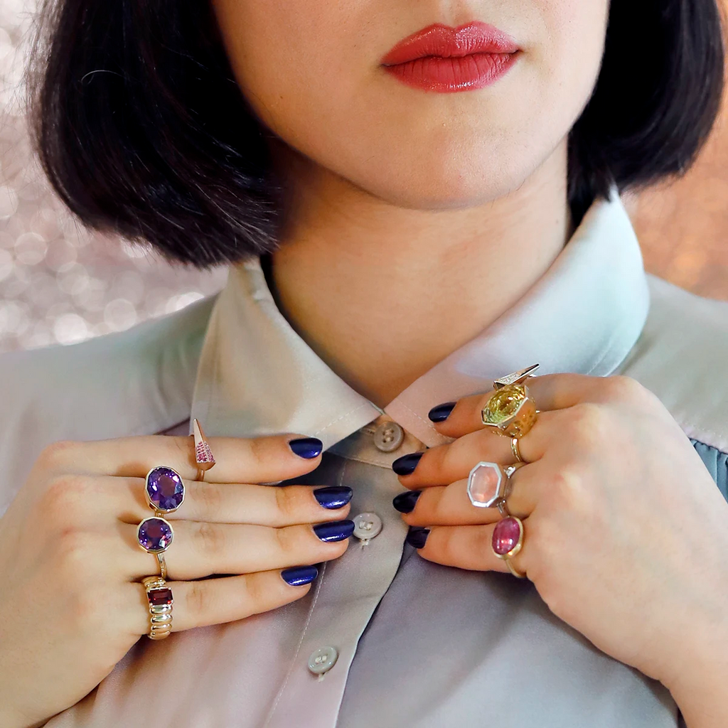 A close-up shot of a young woman's collar and her hands replacing it. We see the lower part of her face, with red lips and bobbed black hair (and a few white hairs). The woman's hands bear 8 absolutely unique and impressive designer rings. There's also a ruby cabochon. There are two modern arrow-shaped rings, one with pavé diamonds and the other with pavé pink sapphires. The last ring worn on the little finger features a central rectangular red garnet and a large swollen gold ring with round shapes.
