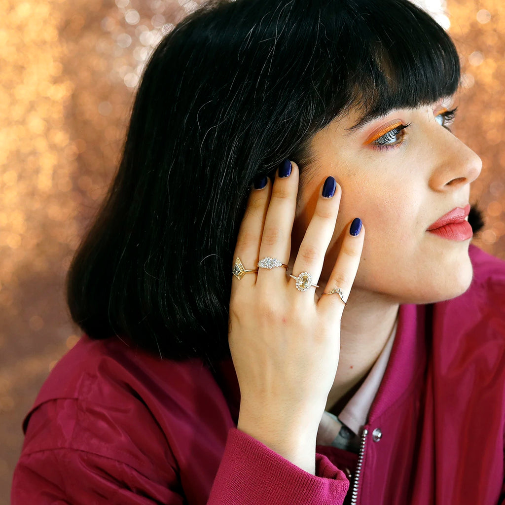 A young woman in a fuchsia blouse, black bobbed hair and orange eyeliner is photographed in front of a pinkish glitter background. She holds her hand to her right cheek. On this hand, she wears 4 pieces of fine jewelry with diamonds and sapphires. These are Canadian designer jewels, all available at Ruby Mardi, a luxury jewelry store in Montreal, Canada.