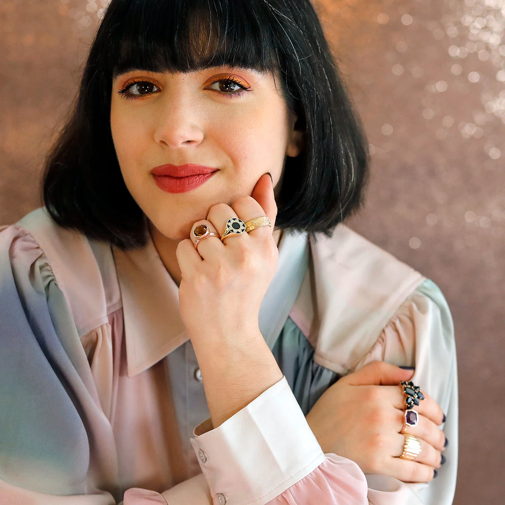 A deep-eyed, light-smiling young woman with bobbed black hair and a pastel blouse is photographed against a background of pink sequins. She wears fine Canadian designer jewelry on both hands. Three pieces are blurred, but three are clearly visible. All are in yellow gold and feature unique details. They are bold pieces of jewelry that empowers and that are unisex jewels.