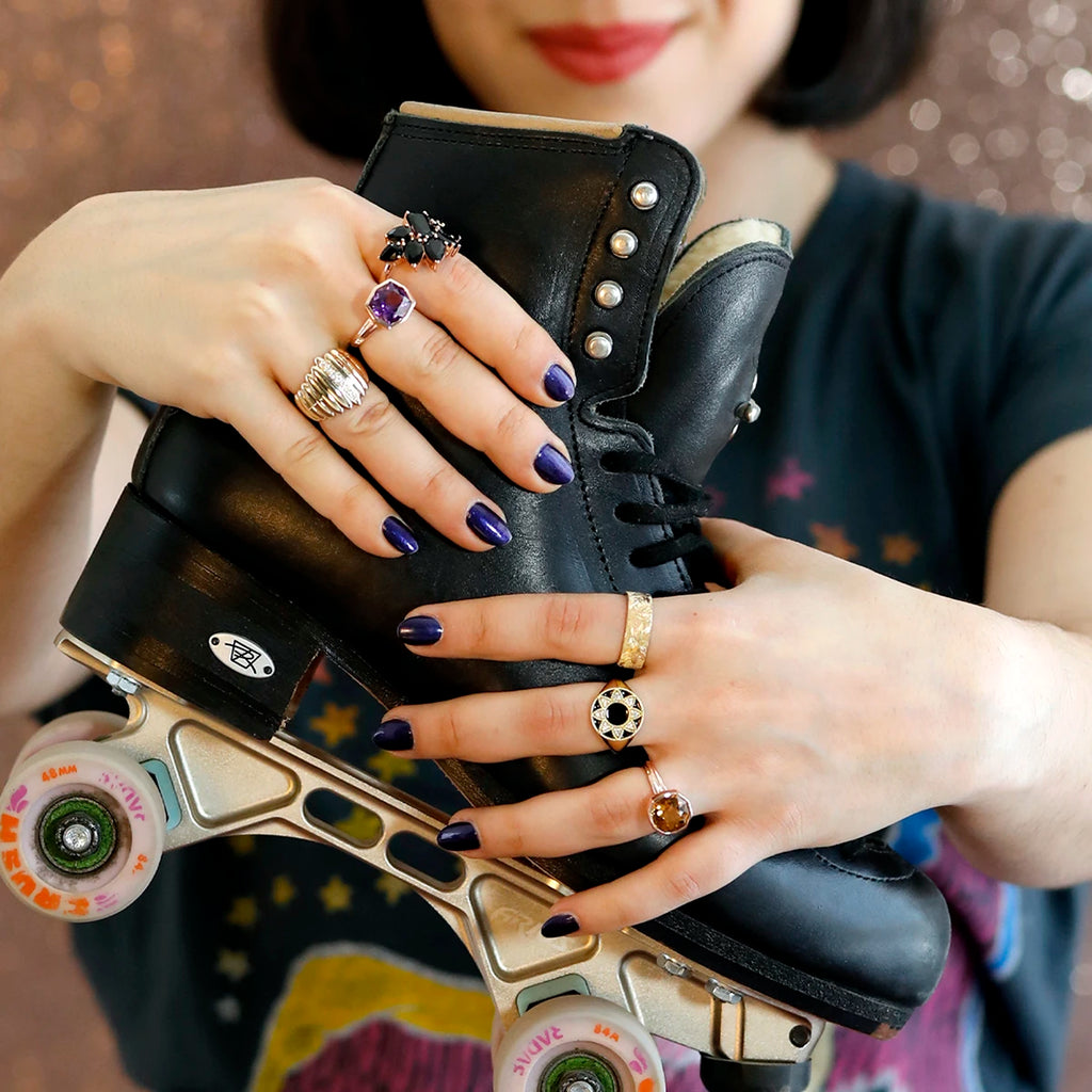 In the foreground of the image: two hands with purple nails hold a black leather roller skate. Blurred behind, a young woman with red lips and black hair bobbed with Pulp Fiction-like energy. The hands carry 6 large, unique Canadian designer rings, available at Canada's coolest jewelry store: Ruby Mardi. The rings are in yellow gold and feature black tourmalines, natural amethysts, citrine, diamonds and finely executed engravings.