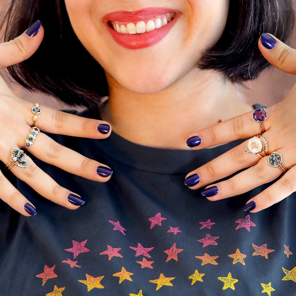 We see the lower face of a smiling girl wearing a dark gray t-shirt with yellow to pink stars, purple nails and several designer rings on her fingers. The rings are unique and all made by Canadian designers. There are rings with ethical sapphires, and cocktail rings with exceptionally large quartz. There are also diamond wedding rings. All these rings are selected and available at Ruby Mardi, the best jewelry store in Canada.
