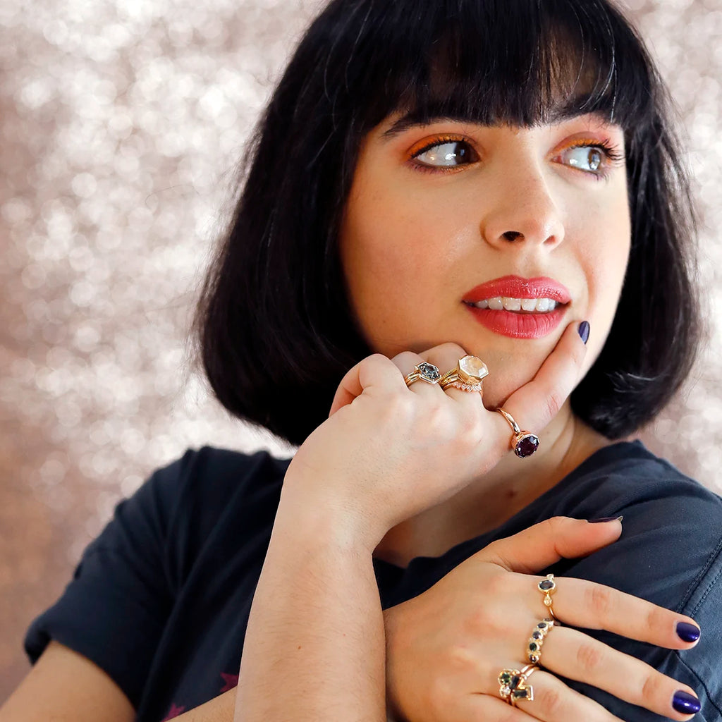 A young woman with red lips and bobbed hair is photographed against a background of pale pink sequins. She looks to her left in a relaxed posture. She wears several handcrafted designer rings on her fingers, eight to be exact. These are precious rings with ethically-crafted natural sapphires, and evening rings with large quartz of exceptional quality. The image exudes a Pulp Fiction-like energy.