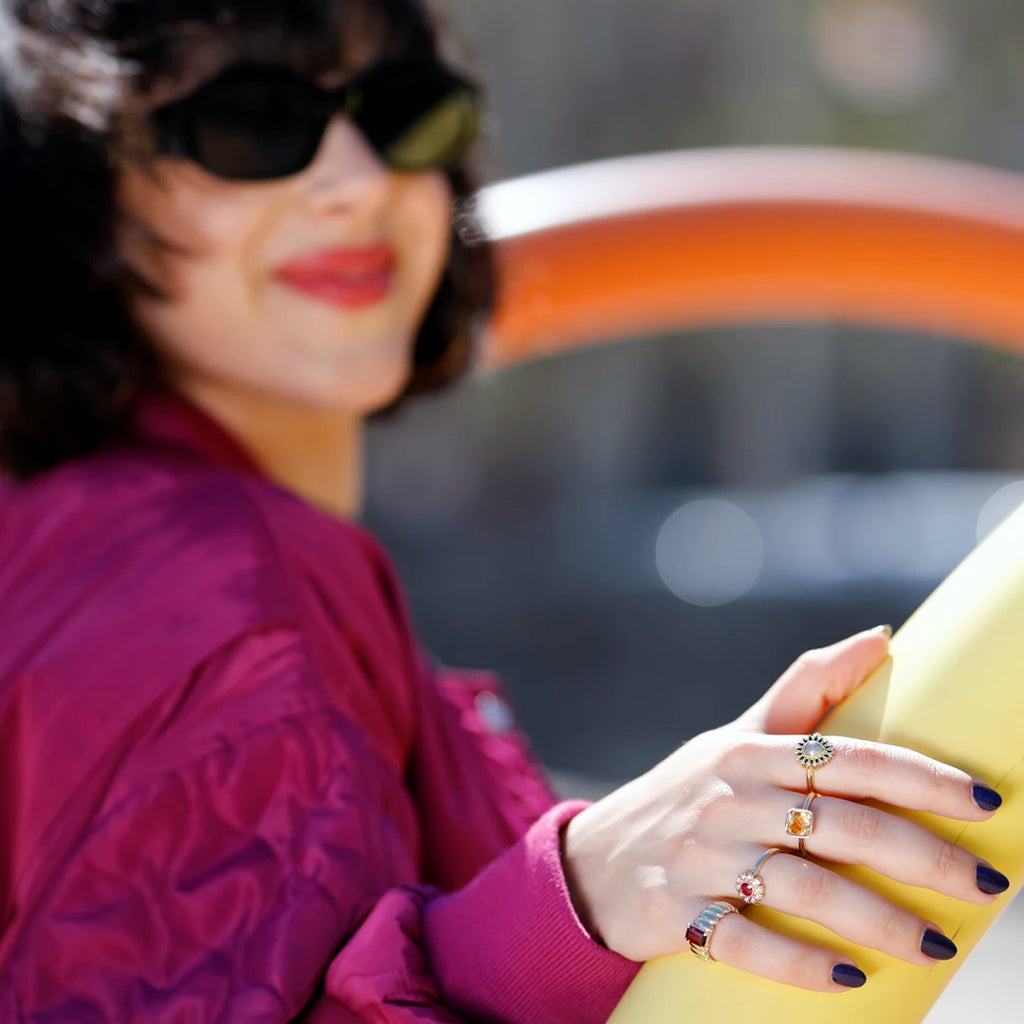 A young woman wearing sunglasses and a fuchsia jacket is photographed outside. Her hand is resting on a yellow children's game, and her hand is clearly visible: she is wearing 4 original rings in different styles, created by 3 different Canadian jewelry designers. All her rings are available at Ruby Mardi fine jewelry. The rings feature these gemstones: red garnet, ruby, citrine and salt-and-pepper diamond.