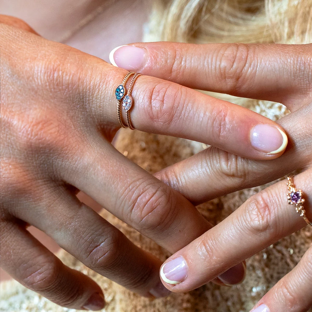 Close-up of a hand wearing two stacked yellow gold rings, each featuring a marquise-shaped diamond cluster — one with blue diamonds and one with white diamonds. Designer jewelry by Ruby Mardi, Montreal, Canada.