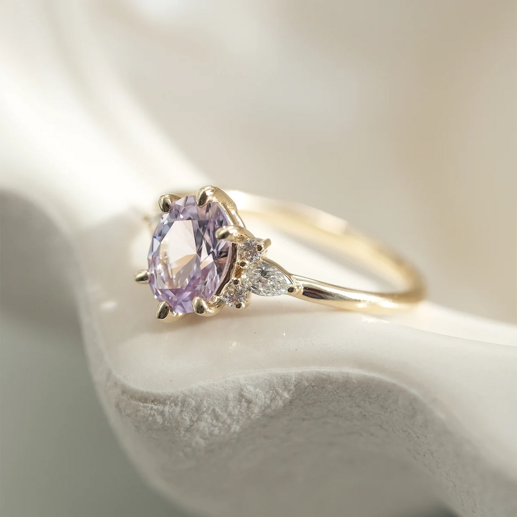 Close-up of lilac sapphire engagement ring with pear-shaped and round diamond accents in 14k yellow gold, resting on a ceramic surface. Handmade in Montreal by Maksym Jewelry for Ruby Mardi.