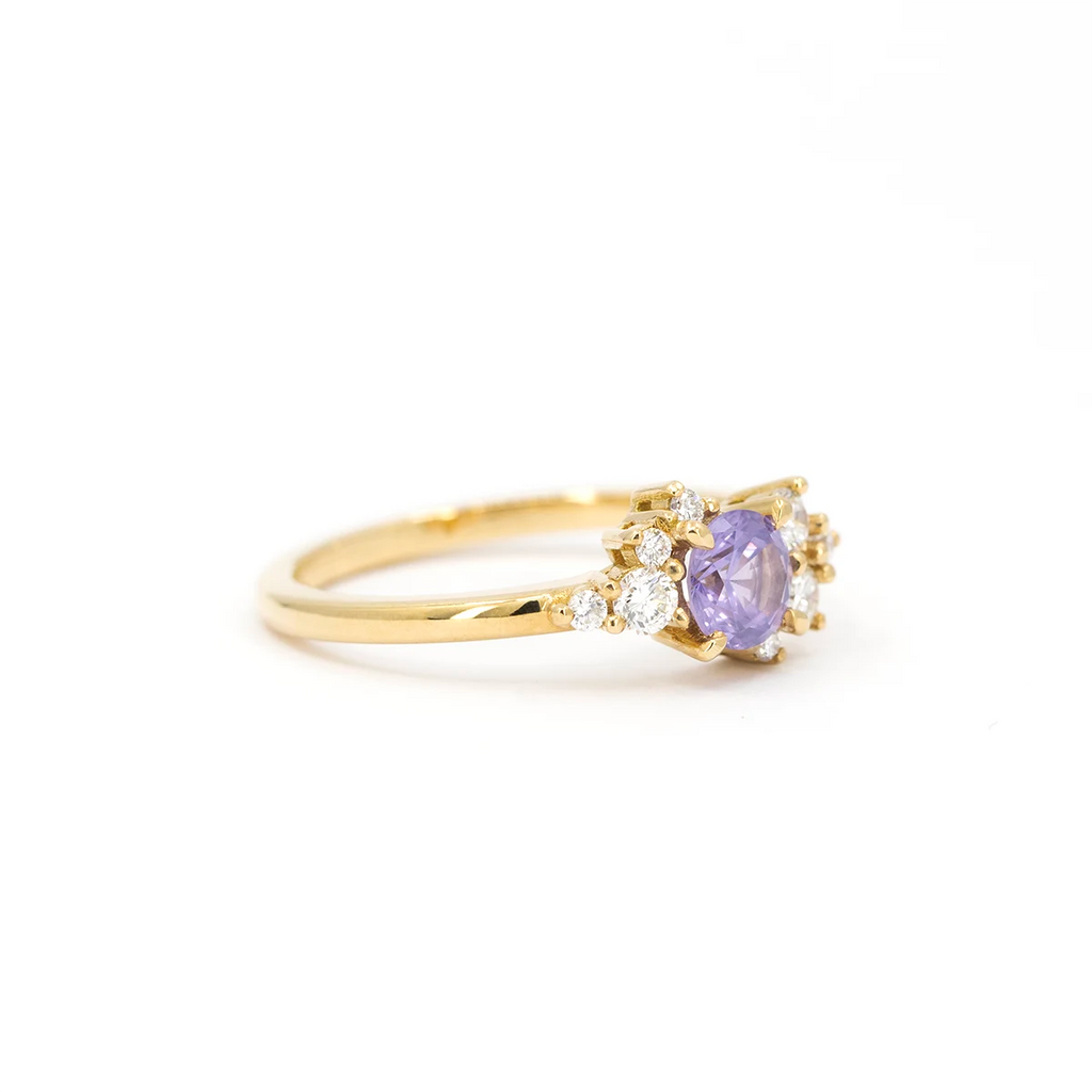 A gold ring with a round purple sapphire center stone, accented by asymmetrically arranged small round white diamonds, displayed at an angle on a white background.