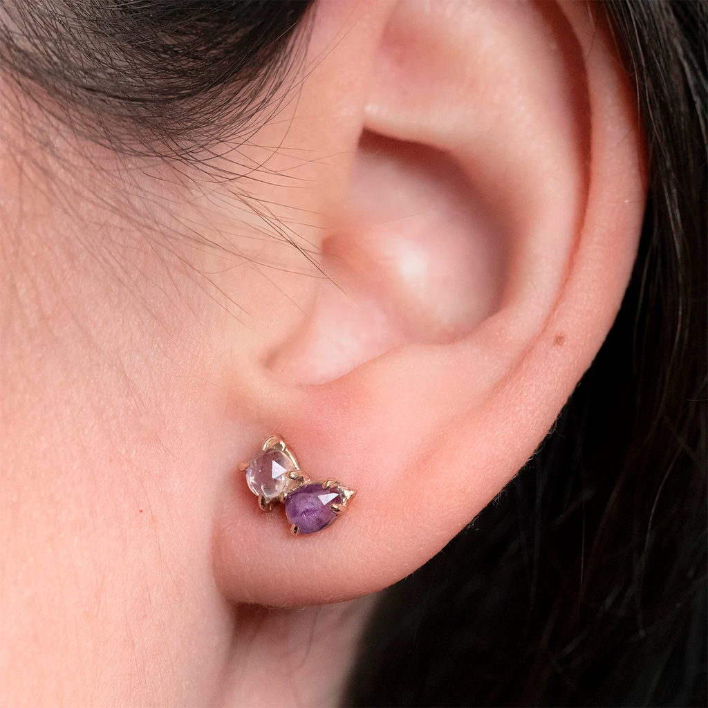 Close-up of a person wearing one-of-a-kind 10k yellow gold stud earrings by Toronto designer Nadia Werchola, featuring natural amethyst and rose-cut moonstone in slightly irregular eagle-claw prongs, shown on the earlobe, available at Ruby Mardi in Montreal.