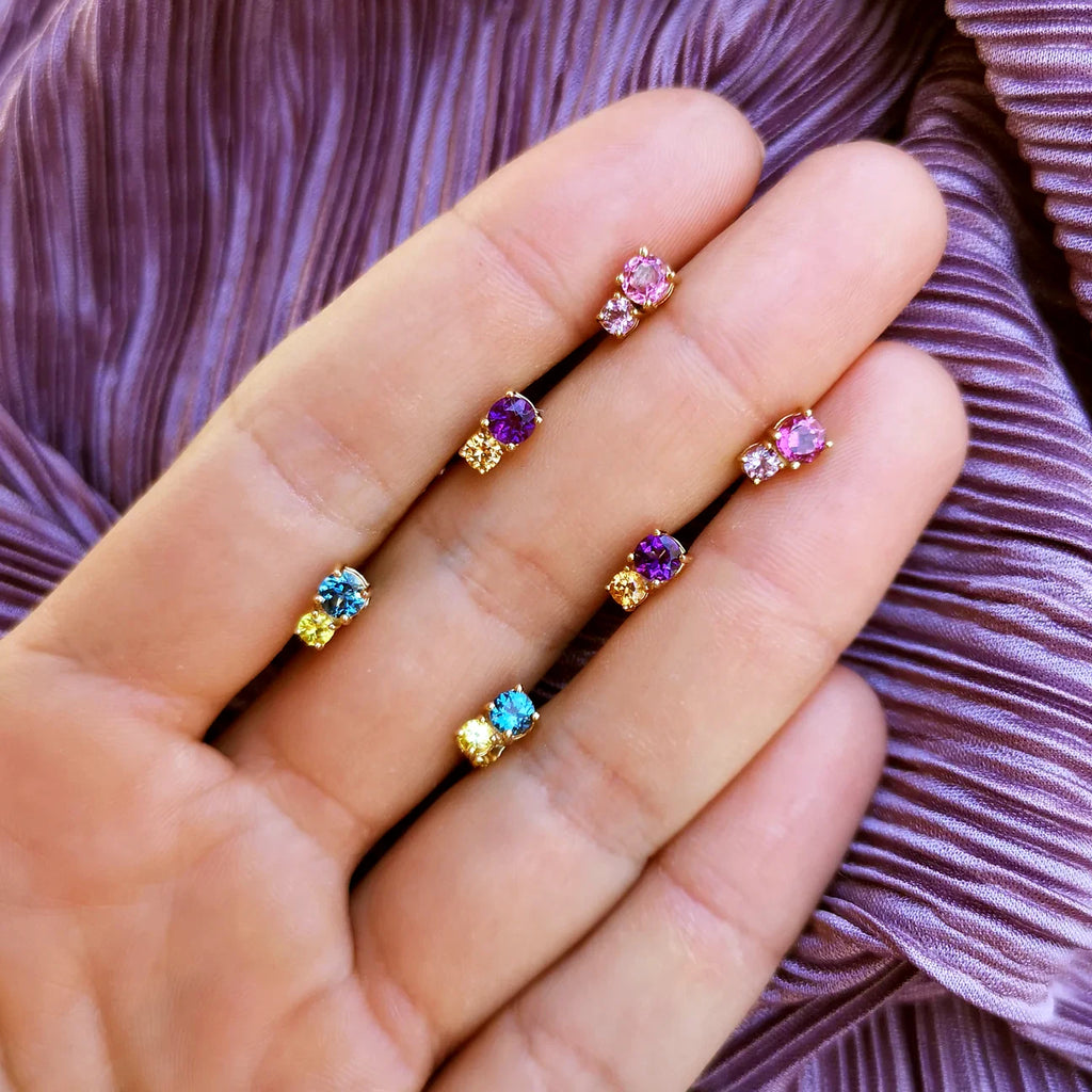 Close-up of six colorful gemstone stud earrings displayed on a hand. Each earring pairs two vibrant stones such as amethyst, blue topaz, citrine, and pink tourmaline, set in yellow gold. Handmade creations by Lico Jewelry, Montreal.