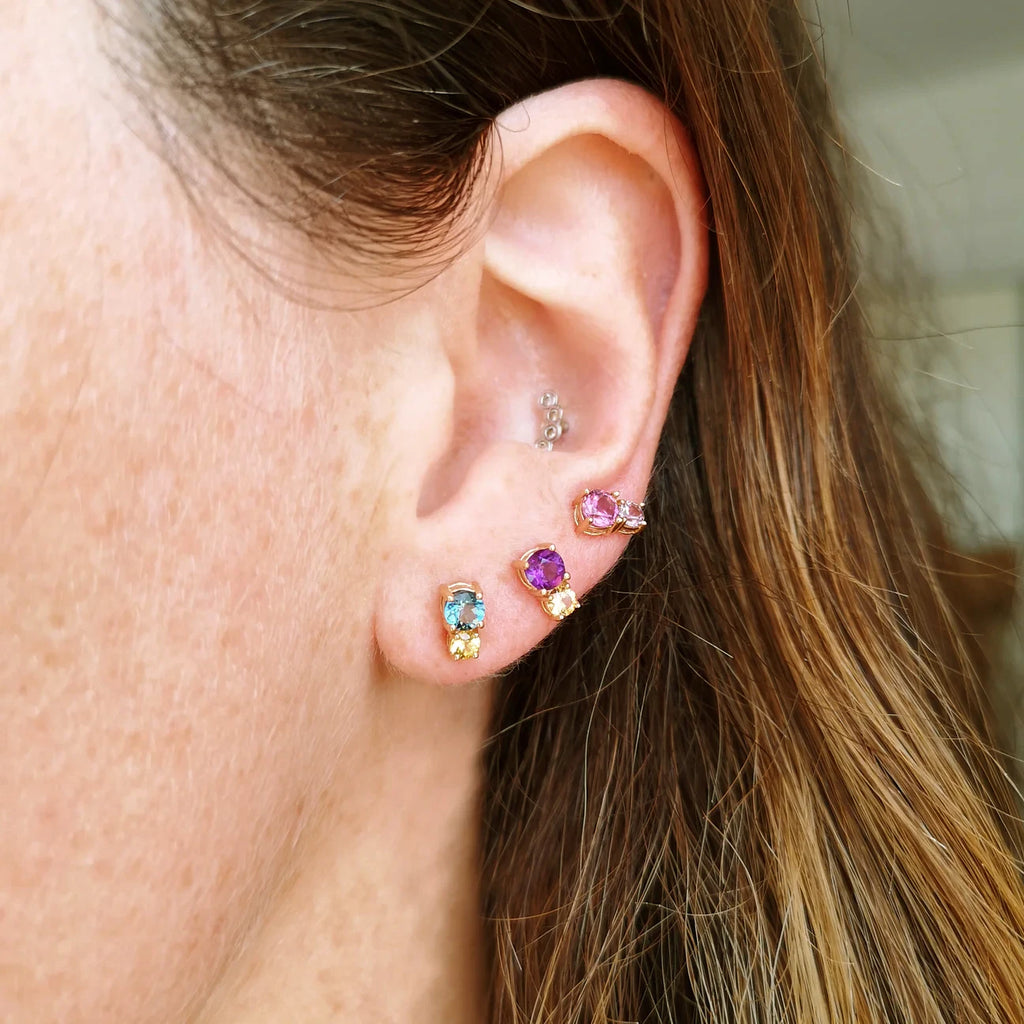 Three colorful gemstone stud earrings worn on the ear — combinations include blue topaz with yellow citrine, amethyst with orange garnet, and pink tourmaline with pale pink sapphire. Playful and elegant jewelry by Lico Jewelry, Montreal.
