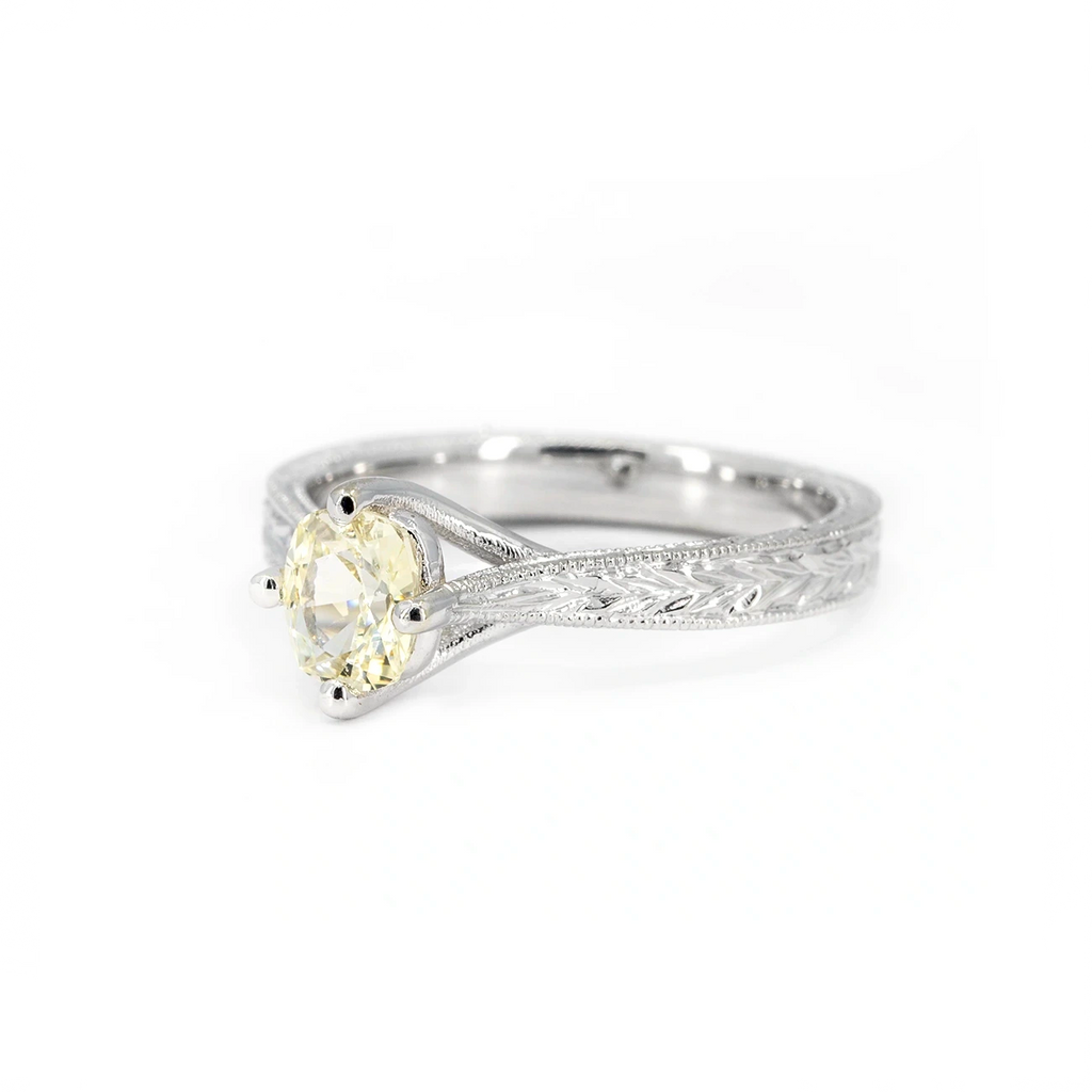 14k white gold solitaire engagement ring featuring a pale yellow oval sapphire set in four prongs, with intricate hand-engraved leaf details on the band. Designed by Deborah Lavery, available at Ruby Mardi, Montreal.