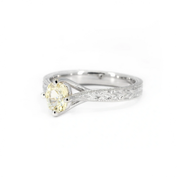 14k white gold solitaire engagement ring featuring a pale yellow oval sapphire set in four prongs, with intricate hand-engraved leaf details on the band. Designed by Deborah Lavery, available at Ruby Mardi, Montreal.