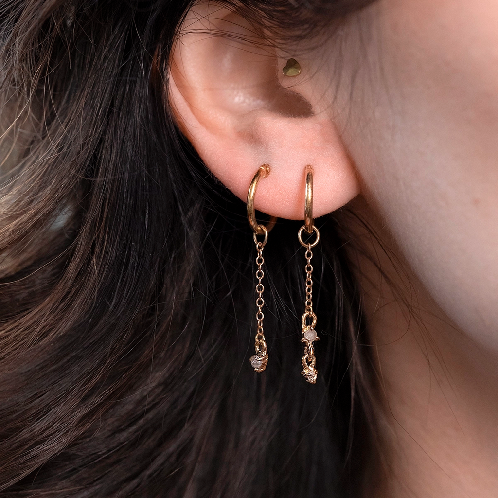 Asymmetrical 10k yellow gold hoop earrings by independent Toronto designer Nadia Werchola, featuring natural rose-cut grey and white diamonds on fine dangling chains, shown worn on the ear with close-up detail, available at Ruby Mardi in Montreal.