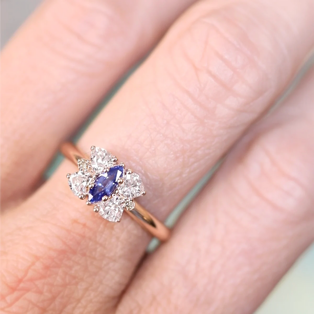 Close-up of the Aurorina ring worn on a hand, featuring a marquise-cut blue sapphire framed by clustered white diamonds in a rose gold band. Handmade and available at Ruby Mardi.