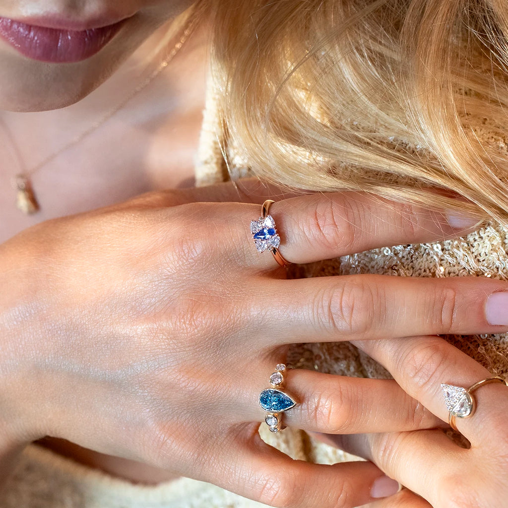 Close-up of a woman’s hands wearing three unique gemstone rings: a rose gold ring with a vivid blue sapphire and diamond cluster, a gold ring with a pear-shaped blue diamond and round diamonds, and a gold ring with a pear-shaped white diamond. Designer jewelry by Ruby Mardi, Montreal, Canada.