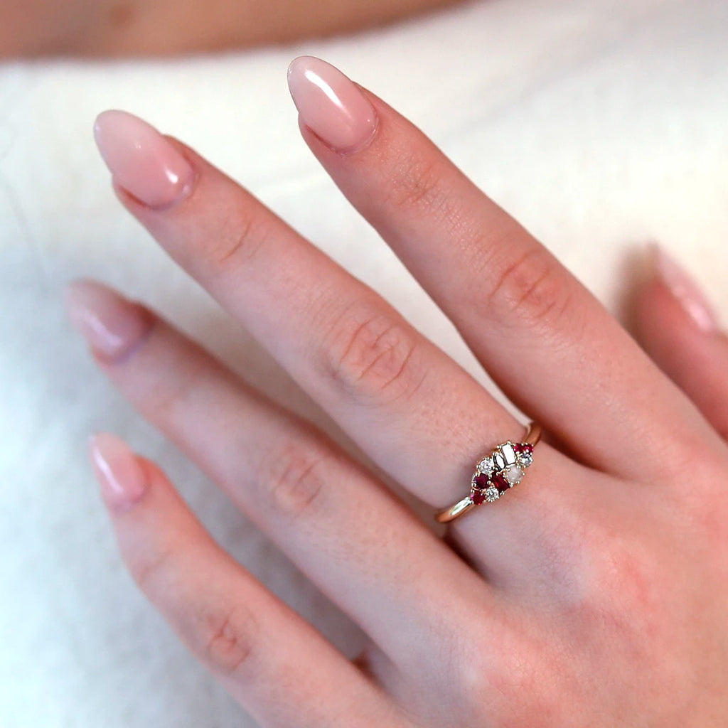 Close-up of a 14k yellow gold cluster ring with rubies, white diamonds, and a salt and pepper diamond, worn on an elegant hand with long fingers and light pink nails. The ring is showcased against a white sweater, emphasizing its luxurious design.