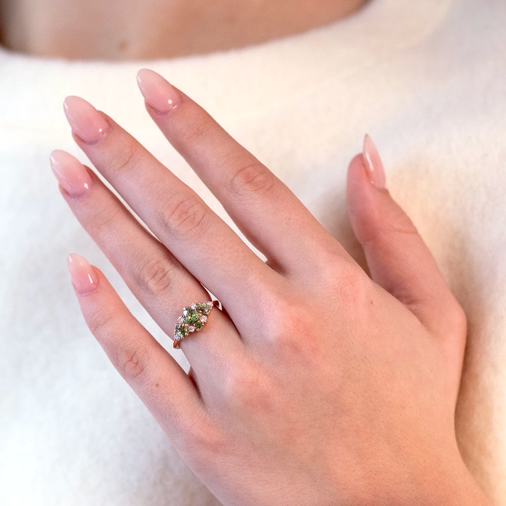 Rose gold ring by Ruby Mardi featuring a cluster of lab-grown diamonds, green sapphires, and demantoid garnets, worn on a hand with long pale pink nails. The hand is posed near the chest, against a light beige knit sweater, highlighting the ring’s vibrant gemstones and elegant design.