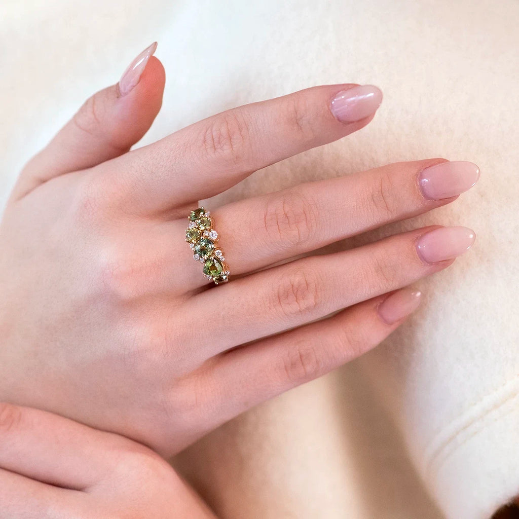 A close-up of a feminine hand adorned with a luxurious green gemstone ring in yellow gold, featuring green sapphires, green garnets, and natural diamonds. This unique creation from Ruby Mardi blends elegance with bold design. Crafted with care in Canada—wearable art at its finest.