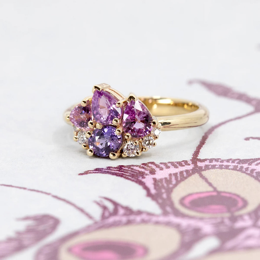 Petal Soufflé by Ruby Mardi placed on an illustrated lilac-toned background. The 14k yellow gold cluster ring combines pink, purple and lilac sapphires with lab-grown diamonds, styled with an artistic backdrop.