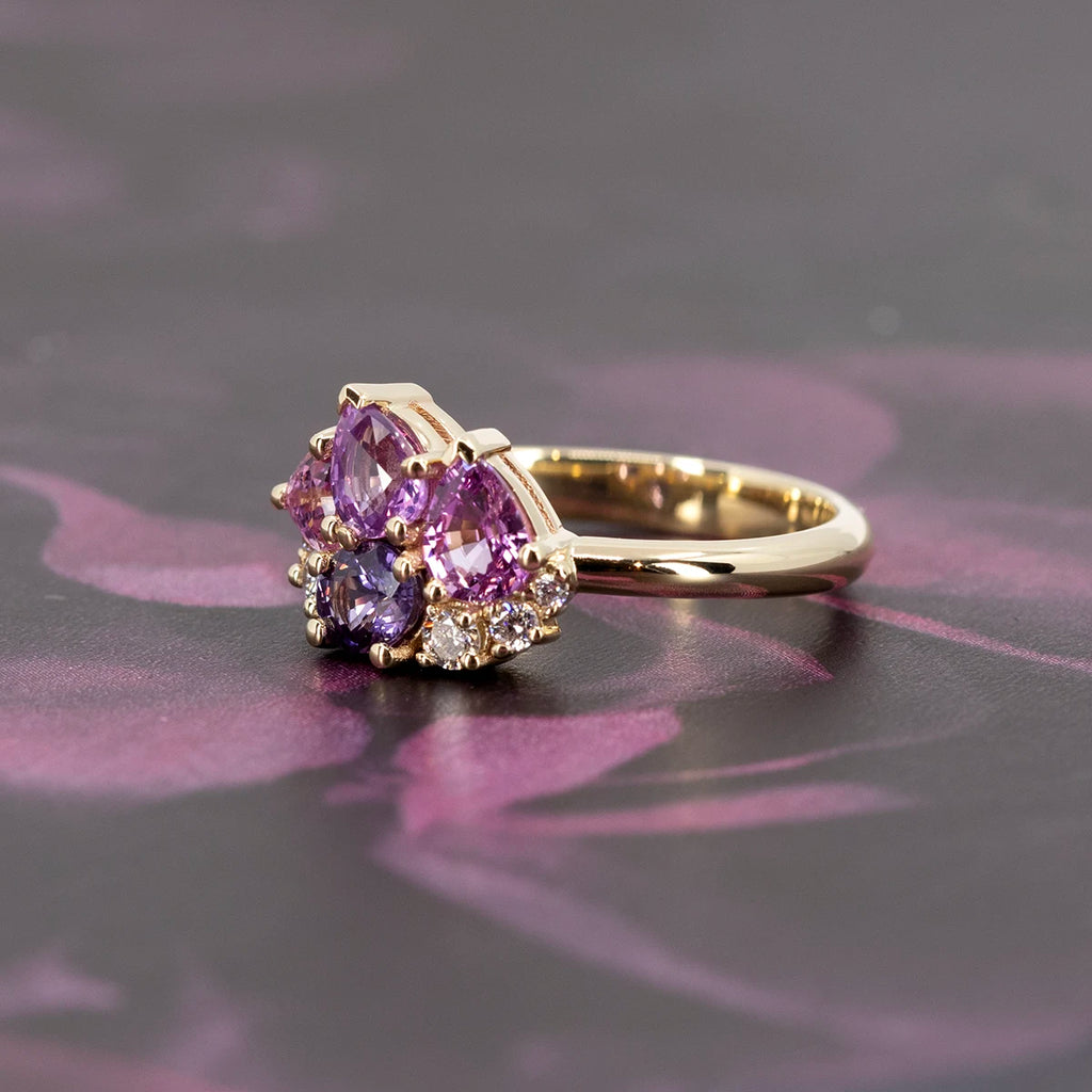 Petal Soufflé cluster ring by Ruby Mardi showcased on a dark floral surface. The pastel sapphires and sparkling lab-grown diamonds in 14k yellow gold contrast beautifully with the deep purple pattern.