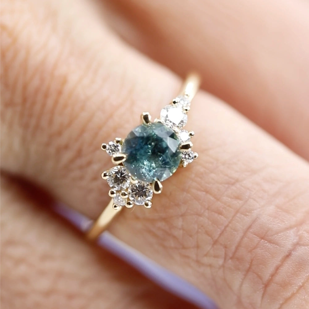 Close-up product photography of a unique engagement ring featuring a stunning round soft blue sapphire and a cluster of natural diamonds. The ring is elegantly worn on a finger, highlighting its asymmetrical design and fine craftsmanship. A one-of-a-kind creation by Justine Quintal, available at Ruby Mardi, Canada's top jewelry store.
